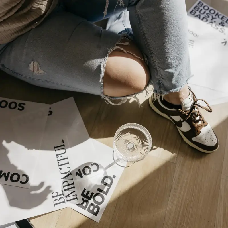 Behind the scenes shot inside the Lenya Creative Studio with Rebel in ripped jeans and sneakers, a champagne glass and papers littering the floor that say "BE IMPACTFUL." "BE BOLD." which are key elements of the Lenya website redesign experience